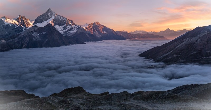 Jagged mountain peaks rising above a sea of clouds at sunrise with pink and orange sky